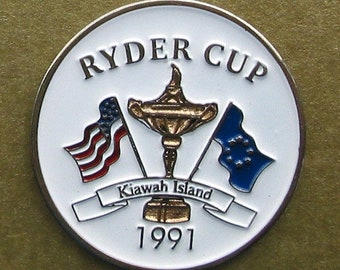 May include: A round Ryder Cup commemorative coin with a white background and gold trim. The coin features the Ryder Cup trophy, American and European flags, and the text "Ryder Cup", "Kawah Island", and "1991".