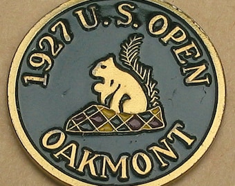May include: A gold-coloured golf ball marker with a black background. The marker features the text "1927 U.S. OPEN OAKMONT" in a gold-coloured font. A cartoon squirrel sits on a checkered surface.