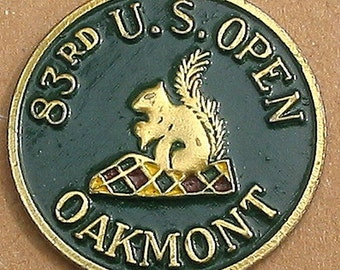 May include: A gold and green golf ball marker with the text "83rd U.S. Open Oakmont" and a gold squirrel sitting on a chequered pattern.