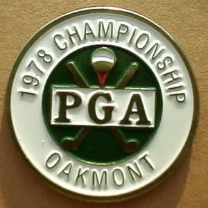 Può includere: Uno spillo rotondo bianco e dorato con il testo "1978 Championship PGA Oakmont" attorno al bordo. Il centro presenta un cerchio verde con due mazze da golf incrociate e una palla da golf bianca.