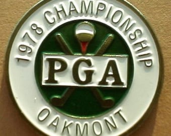 Puede incluir: Un pin redondo blanco y dorado con el texto "1978 Championship PGA Oakmont" alrededor del borde. El centro presenta un círculo verde con dos palos de golf cruzados y una bola de golf blanca.
