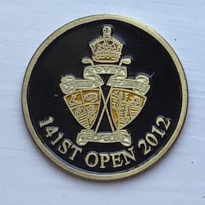 Op de afbeelding: Een zwarte en gouden golfbalmarker met de tekst "141st Open 2012" en een wapenschild met een kroon, gekruiste golfclubs en de woorden "Royal St Andrews Golf Club".