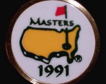 Puede incluir: Una bola de golf blanca con un borde dorado. La bola presenta un contorno verde y amarillo de los Estados Unidos con una bandera de golf y un agujero. El texto "Masters" y "1991" está impreso en verde.