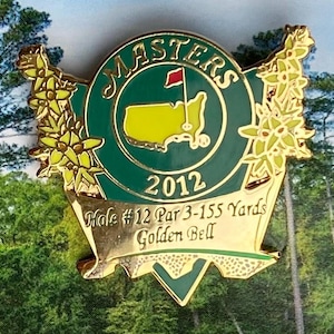Op de afbeelding: Een goudkleurige Masters golfspeld met een groen en geel embleem. De speld toont het Masters-logo, het jaar 2012 en tekst met Hole #12, Par 3-155 Yards en Golden Bell. De speld is versierd met bloemaccenten.