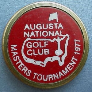 May include: A red and white golf ball marker with the text "Augusta National Golf Club Masters Tournament 1977". The marker features a white outline of the United States with a golf course inside.