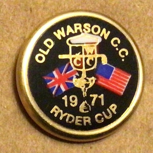 May include: A gold-colored golf ball marker with a black background. The marker features the text "OLD WARSON C.C." and "1971 RYDER CUP" along with a graphic of two flags, a golf club, and a golf ball.