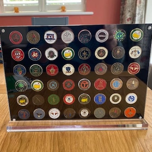 Puede incluir: Una colección de pines y insignias redondas vintage exhibidas en un soporte acrílico transparente. Los pines presentan varios colores y diseños, incluyendo logotipos, emblemas y texto. La exhibición está sobre una mesa de madera.