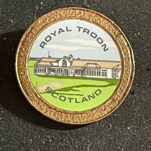May include: A gold-coloured round pin with a white background featuring a drawing of a building with the text "Royal Troon Golf Course Scotland".