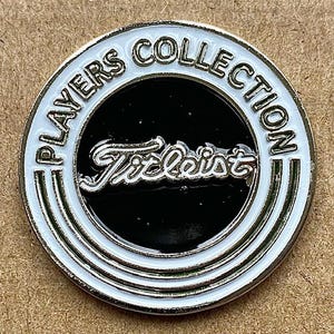 Könnte beinhalten: Ein kreisförmiger Golfballmarker mit dem Text "PLAYERS COLLECTION" am Rand. In der Mitte befindet sich das Wort "Titleist" in Schreibschrift auf schwarzem Hintergrund. Der Marker hat ein weißes und silbernes Farbschema.