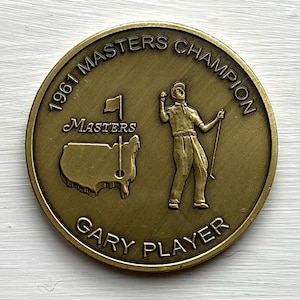 May include: A gold-colored coin commemorating the 1961 Masters Champion, Gary Player. The coin features a golfer raising a club in the air, a map of the United States with the word "Masters" above it, and the text "1961 Masters Champion Gary Player".