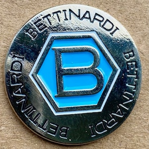 Puede incluir: Un marcador de bola de golf de color plateado con el texto "Bettinardi" alrededor del borde y una "B" azul dentro de un hexágono blanco en el centro.