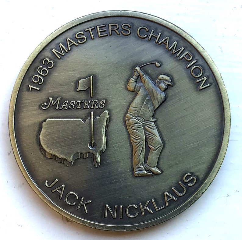 K&ouml;nnte beinhalten: Eine goldfarbene M&uuml;nze zur Erinnerung an Jack Nicklaus' Sieg beim Masters 1963. Die M&uuml;nze zeigt einen Golfer beim Abschlag, eine Karte der Vereinigten Staaten und den Text "1963 Masters Champion Jack Nicklaus".