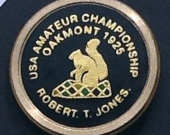 May include: A gold-coloured metal pin with a black background. The pin features a squirrel design with the text "USA Amateur Championship Oakmont 1925 Robert T. Jones."