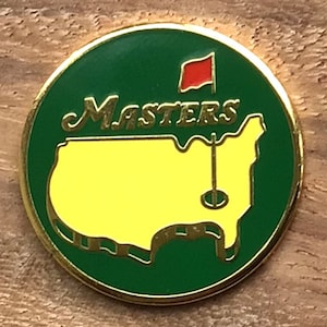 Pode incluir: Uma bola de golfe verde e amarela com as palavras "Masters" e uma representação dos Estados Unidos.