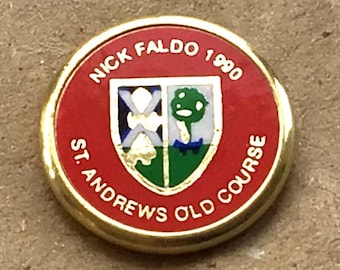 Puede incluir: Un marcador de bola de golf rojo y dorado con el texto "Nick Faldo 1990 St. Andrews Old Course" y un escudo con una cruz blanca y un árbol verde.