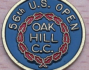 Op de afbeelding: Een blauwe en gouden golfbalmarker met de tekst "56th U.S. OPEN OAK HILL C.C." omringd door een rode en gouden krans.