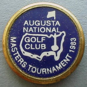 May include: A round, gold-toned pin with a blue center featuring white text and a golf course outline. The text reads "AUGUSTA NATIONAL GOLF CLUB MASTERS TOURNAMENT 1983". A small white flag is at the top.