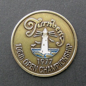 May include: A gold-colored metal coin with a white lighthouse and blue water design. The text "Turnberry 106th Open Championship 1977" is inscribed around the edge.
