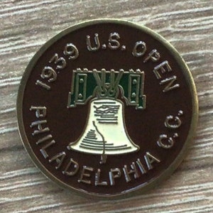Puede incluir: Un marcador de bola de golf de metal marrón y dorado con el texto "1939 U.S. OPEN PHILADELPHIA C.C." y una representación de la Campana de la Libertad.