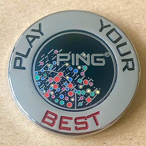 May include: A circular silver-toned metal golf ball marker featuring the text PLAY YOUR BEST in raised lettering. The center has a black background with the PING logo and a graphic pattern of small, colorful circles in red, blue, and green.