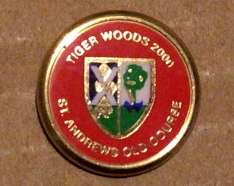 Puede incluir: Un marcador de bola de golf rojo y dorado con el texto "Tiger Woods 2000 St. Andrews Old Course" alrededor del borde. El centro presenta un escudo verde, blanco y azul.