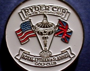 May include: A white and silver Ryder Cup commemorative coin. The coin features the text "RYDER CUP" above a trophy and the flags of the United States and Great Britain. The text "ROYAL LYTHAM & ST. ANNES GOLF CLUB" is also present.
