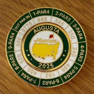 May include: A gold-colored golf ball marker with the Augusta Masters logo, a yellow map, and the text "2025." The marker also displays hole numbers, par values, and yardages, including "1-PAR4 445" and "2-PAR5 585."