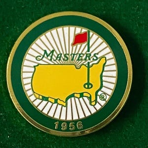 May include: A round, gold-rimmed pin with a green border. The center features a yellow map of the U.S. with a red flag, the word "MASTERS" in green, and the year "1956" at the bottom. The background has white rays.