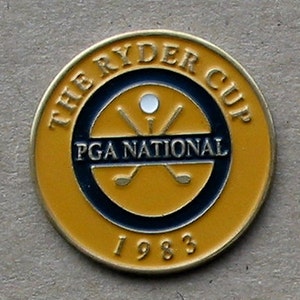 May include: A gold-colored metal coin with a black and yellow design. The coin features the text "The Ryder Cup" and "PGA National" with a golf ball and crossed golf clubs in the center. The year "1983" is printed at the bottom.