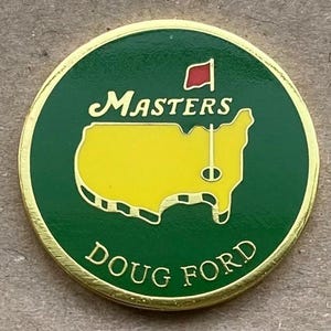May include: A round, gold-edged golf ball marker with a green background. The marker features the word "Masters" in script above a yellow outline of a golf course and the words "Doug Ford" below.