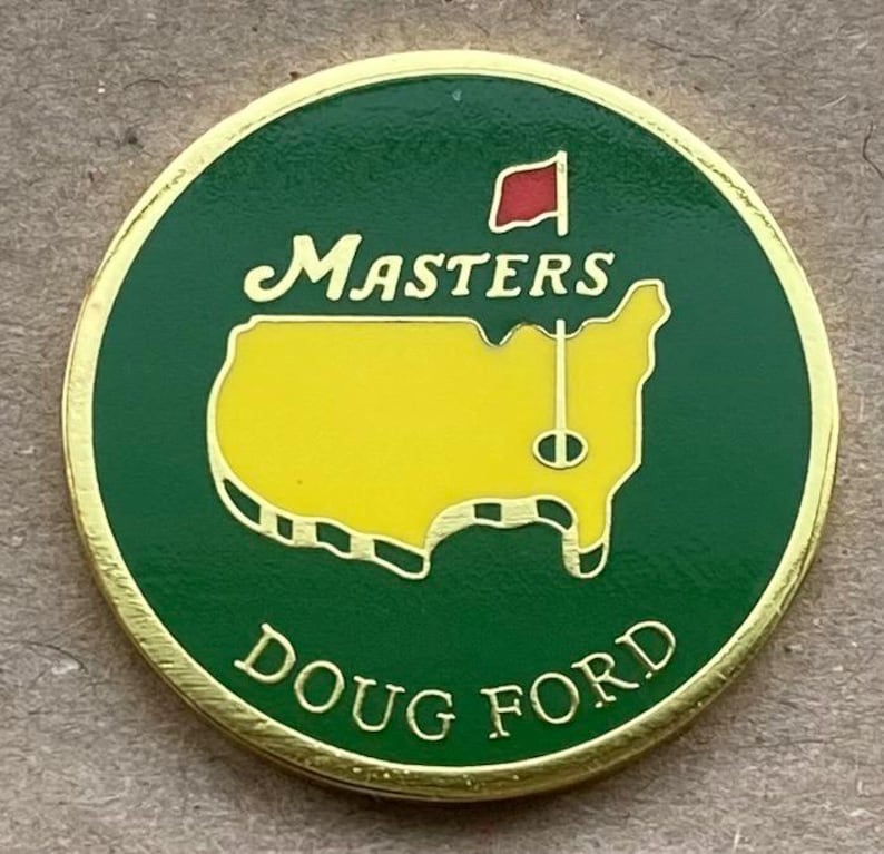 May include: A round, gold-edged golf ball marker with a green background. The marker features the word "Masters" in script above a yellow outline of a golf course and the words "Doug Ford" below.