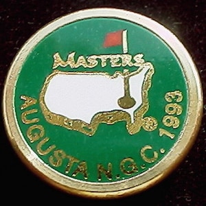 May include: A gold-coloured golf ball with a green background and a white outline of the United States. The text "Masters" is written in gold above the outline and "Augusta N.G.C. 1993" is written in gold around the outline.
