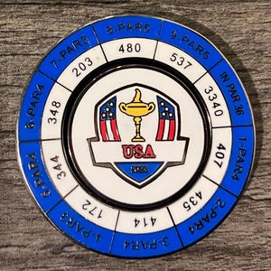 May include: A white and blue circular golf scorecard with a red, white, and blue USA logo in the center. The scorecard has a golf trophy in the center of the logo. The scorecard has numbers and the words "PAR" and "IN PAR 36" around the edge.