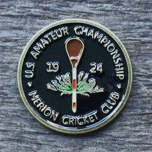 May include: A black and gold metal coin with the text "US Amateur Championship Merion Cricket Club" around the edge. The centre of the coin features a brown cricket bat, a green and white cricket wicket, and the numbers "19" and "24".
