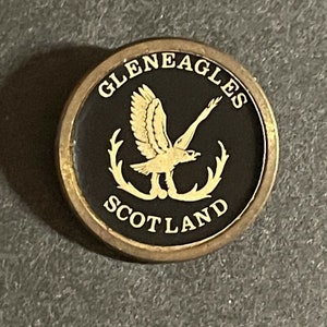 May include: A gold-colored metal pin with a black background. The pin features a gold eagle with outstretched wings and a wreath of antlers. The text "GLENEAGLES SCOTLAND" is written in a gold font around the eagle.