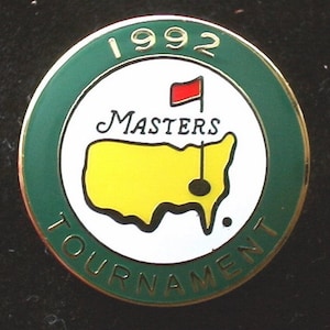 May include: A green and gold circular pin with a white centre. The centre features a yellow outline of the United States with a red flag and a golf hole. The text "1992 Masters Tournament" is printed around the edge.