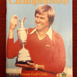 Op de afbeelding: Een rood-wit programma voor het Open Golf Championship dat in juli 1982 in de Royal Troon Golf Club werd gehouden. Het programma bevat een foto van een golfer die de kampioensbeker vasthoudt. De tekst "Official Programme £1.50" staat onderaan het programma gedrukt.