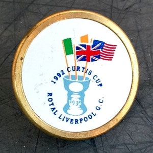 Könnte beinhalten: Ein runder weißer und goldener Pin mit dem Text "1992 Curtis Cup Royal Liverpool G.C." und einer blauen Trophäe mit drei Flaggen: grün, orange und rot, weiß und blau.