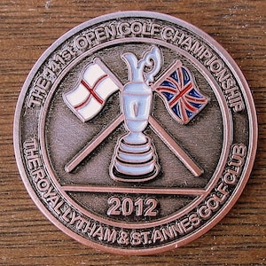 Op de afbeelding: Een bronzen herdenkingsmunt met de tekst "The 41st Open Golf Championship The Royal Lytham & St. Annes Golf Club 2012" rond de rand. Het midden van de munt heeft een witte en blauwe golf trofee met gekruiste vlaggen van Engeland en het Verenigd Koninkrijk.