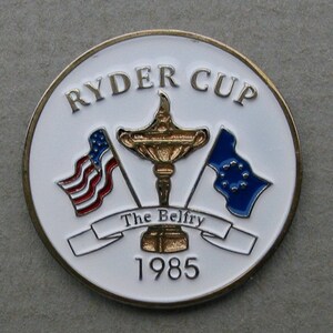 May include: A white, round pin with a gold border. The pin features a gold trophy with a red, white, and blue American flag and a blue European flag on either side. The text "Ryder Cup" is at the top of the pin, and "The Belfry 1985" is at the bottom.