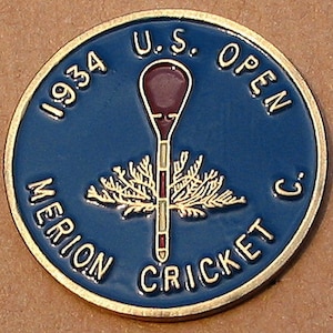 Könnte beinhalten: Eine runde, blau-goldene Münze mit der Aufschrift "1934 U.S. OPEN MERION CRICKET C.". Die Münze zeigt ein weinrotes und weißes Golfschläger-Design mit einem goldfarbenen, blattartigen Emblem.