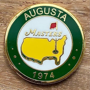 May include: A round, gold-rimmed pin with a green border and the word "AUGUSTA" at the top. The center features a white background with "Masters" in script, a red flag, and a yellow map. The year "1974" is at the bottom.