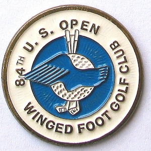 Op de afbeelding: Een witte en gouden golfbalmarker met de tekst "84th U.S. Open Winged Foot Golf Club" rond de rand. Het midden heeft een blauwe cirkel met een witte golfbal en een blauwe gevleugelde voet.
