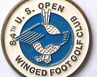 Op de afbeelding: Een witte en gouden golfbalmarker met de tekst "84th U.S. Open Winged Foot Golf Club" rond de rand. Het midden heeft een blauwe cirkel met een witte golfbal en een blauwe gevleugelde voet.