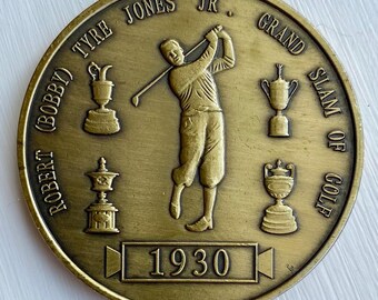 May include: A gold-colored coin commemorating Bobby Jones Jr.'s 1930 Grand Slam of Golf. The coin features a depiction of Jones swinging a golf club, with four trophies representing the four major championships he won that year.