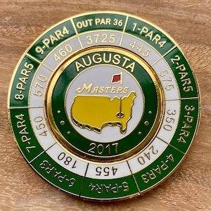 May include: A gold and green golf ball marker with the words "Augusta Masters 2017" and a map of the United States. The marker has a white background with black numbers and the words "Par 4" and "Par 5" around the edge.