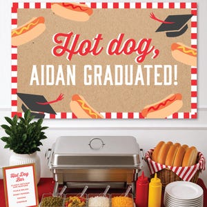 Puede incluir: Una presentación de barra de hot dogs con un letrero que dice "Hot dog, Aidan Graduated!" en rojo y blanco. La escena incluye un calienta platos, condimentos, aderezos y una cesta de panecillos. Un pequeño letrero enumera los aderezos.