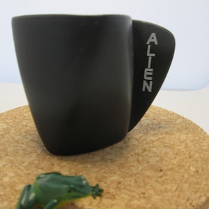 May include: Black ceramic mug with a curved handle and the word "ALIEN" printed on the side. The mug is sitting on a cork coaster.