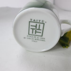 May include: White ceramic mug with a green design and the text "Tait&ugrave;&reg;" and "Foglie &copy; 1996 by Emilio Bergamini, Italy" on the bottom.