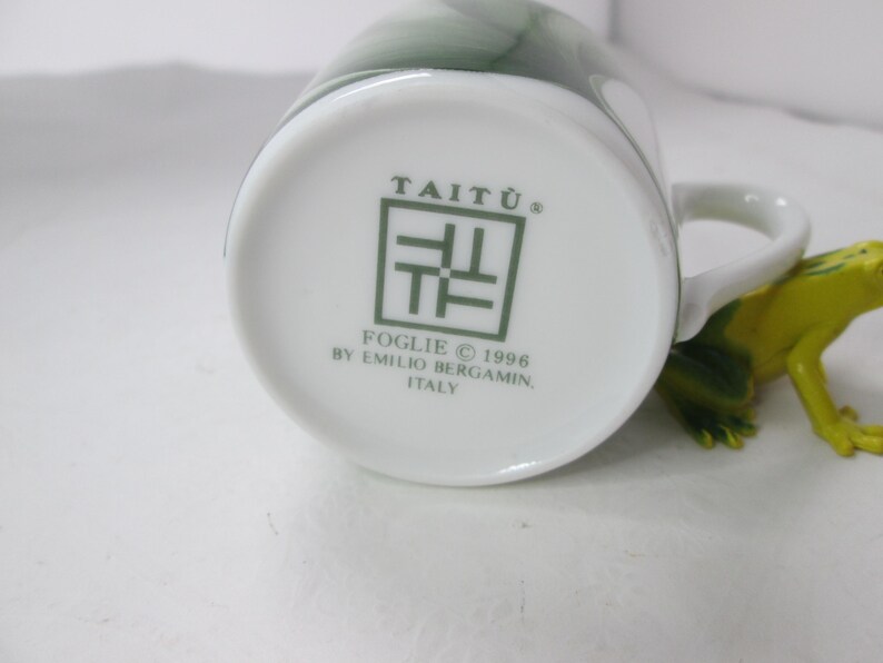 May include: White ceramic mug with a green design and the text "Tait&ugrave;&reg;" and "Foglie &copy; 1996 by Emilio Bergamini, Italy" on the bottom.